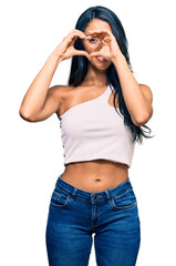 Beautiful hispanic woman wearing casual clothes doing heart shape with hand and fingers smiling looking through sign