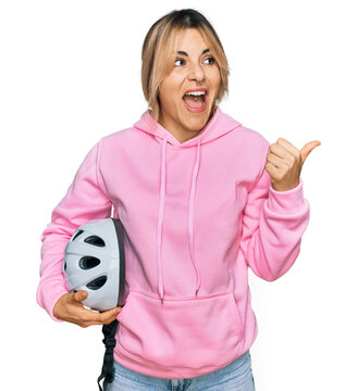 Young caucasian woman holding bike helmet pointing thumb up to the side smiling happy with open mouth