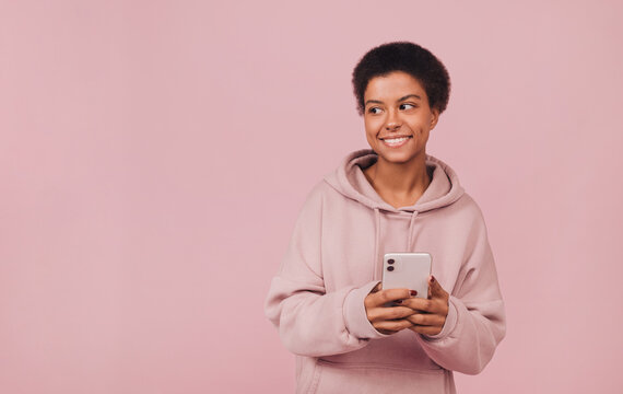 Happy Attractive Black Girl Using Smartphone. Portrait With Copy Space. Cheerful Smiling Young African Woman Wearing Casual Hoodie With Cell Phone While Standing Over Pink Background