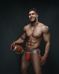 Fototapeta na wymiar Cheerful handsome man with ball in studio. Naked smiling guy in orange jocks underwear with american football ball in his hand.