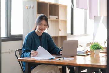 Happy casual young Asian woman working in a home or small office with using a laptop and documenting reports on a desk as a freelancer.