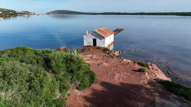 Old boat shed on the banks of Fornells Bay in Menorca Spain with busy pier and birds claiming home.
