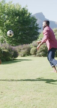 Vertical video of african american father and son playing football in garden, in slow motion