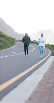 Vertical video of senior biracial couple running on road in mountains, in slow motion