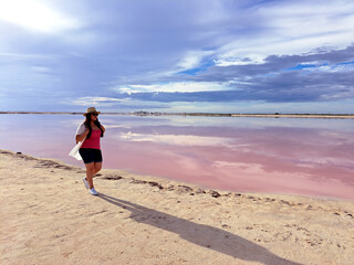 Latin adult woman with shorts, pink shirt, hat and sunglasses walks on the sand next to the pink colored lagoon with a high concentration of salt, Las Coloradas in Yucatan Mexico