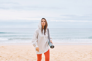 Sweet young woman in white hoodie, walking with yoga mat, on seashore in early hours of morning