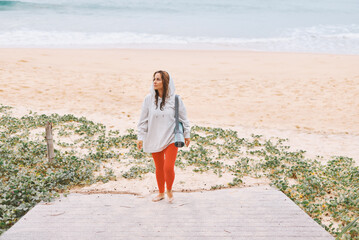 Sweet young woman in white hoodie, with yoga mat on shoulder, came to the beach to do yoga