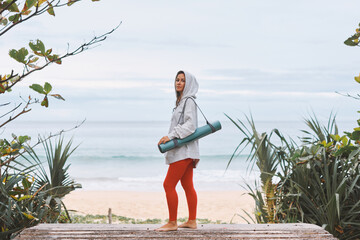 Young woman with yoga mat,on shoulder standing on seashore in early morning, going to do yoga
