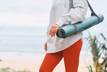 Unrecognizable woman with yoga mat on shoulder, strolling along seashore in early hours of morning