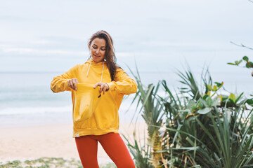 Attractive young girl in yellow hoodie, yellow hat standing on seashore in a relaxed state