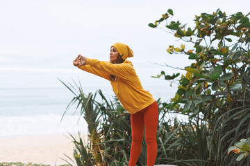 Back stretching Young athletic woman in yellow hoodie, yellow hat doing yoga exercises in front of sea