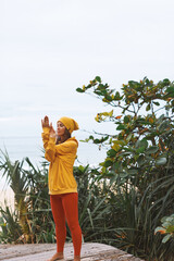 Balancing Young athletic woman in yellow hoodie, yellow hat doing yoga exercises in front of sea