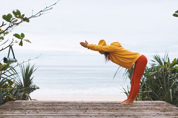 Balancing Young athletic woman in yellow hoodie, yellow hat doing yoga exercises in front of sea - 576553299