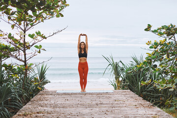 Attractive young athletic woman in black sport bra, red pants doing yoga exercises in front of sea