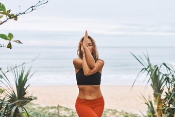 Eagle Pose Attractive athletic woman in black bra, red pants doing yoga exercises in front of sea