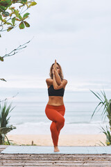 Eagle Pose Young athletic woman in black sport bra, red pants doing yoga exercises in front of sea