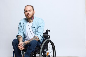 A man in a wheelchair looks thoughtfully at the camera, copy space, with tattoos on his arms sits...