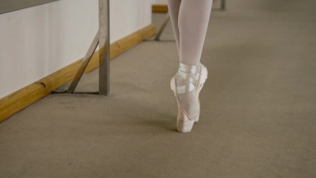 Professional ballerina in training suit dancing on tiptoe in pointe shoes at ballet studio. Ballet dancer prepares for performance. Woman doing classic ballet exercises. Ballet dance school. Zoom out.