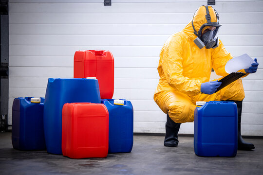Factory worker wearing protection suit and gas mask checking inventory of chemicals inside warehouse.
