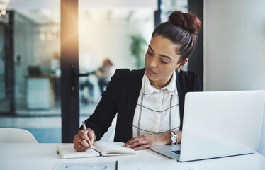 Success is a consistent, daily effort. Shot of a young businesswoman making notes at her desk in a...