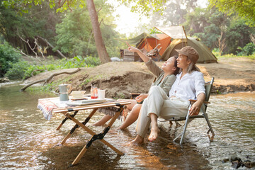Pleased happy mother and daughter reading a book and using laptop while relaxing on the deck chairs in the river, sit near a camp and tent, drink coffee in a pine forest. Camping, recreation, hiking.
