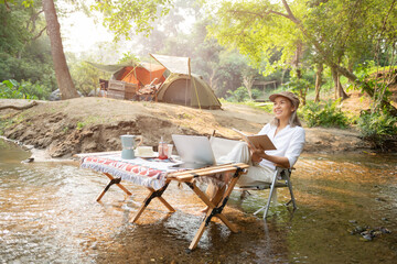 Asian woman travel and camping alone reading a book and using laptop while relaxing on the deck chairs in the river at natural park in Thailand. Recreation and journey outdoor activity lifestyle.