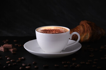 White cup with cappuccino on a dark background, coffee beans on the table. Cappuccino with croissant on a black background.