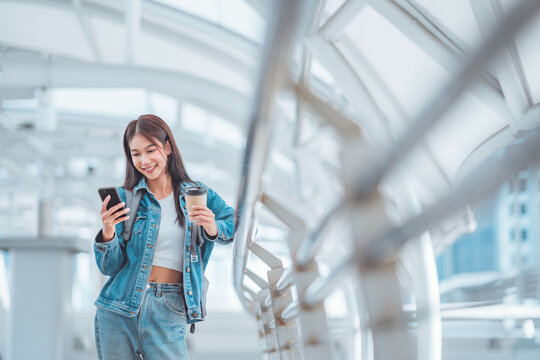 Happy Young Asian Woman Using Social Media On Mobile Phone While Holding Coffee Cup In A Urban City. Copy Space.