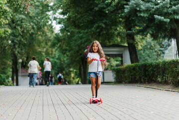A beautiful smiling girl rides a scooter along the path of an amusement park on a summer day. Seasonal children's active sport.