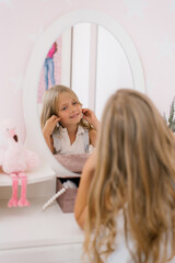 Happy child girl of seven years old puts on earrings in front of the mirror