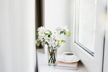 White cup of coffee on a white plate, a stack of books and a vase of flowers on the windowsill Cozy Easter, spring still life.