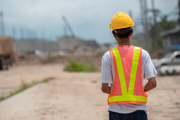 Asian male engineering worker wearing hard hat and protective vest standing back and look forward at construction site.