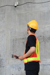 Asian engineering worker wearing hard hat and protective vest using tablet to check in closed-circuit camera in construction site.