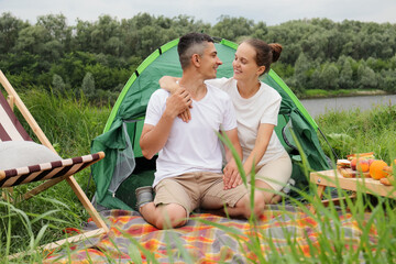 Outdoor shot of happy man and a woman sitting near the tent at the lake or river, having picnic in open air, couple looking at each other and hugging, having fun together.