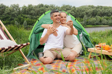 Portrait of man and a woman sitting near the tent at the lake or river on blanket, having picnic, making selfie on mobile phone, smiling to phone camera, expressing happiness.