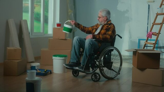 An elderly disabled man moves in a wheelchair takes a bucket of paint, roller and plans to paint the walls. A pensioner plans repairs and wall decoration. Room with window, ladder, cardboard boxes.