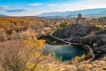 Fototapeta na wymiar Autumn colorful picture of Cetina river source water hole and Orthodox church on the hill view, Dalmatian Zagora region of Croatia. Cetina River Spring. Fall colors leafs on trees. Water landscape.
