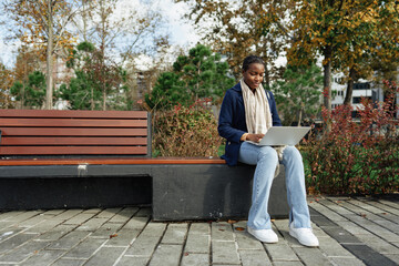 Fototapeta premium Young african businesswoman using laptop while sitting on the bench in the city