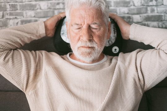 Handsome senior man sitting on the couch with a massage pillow behind his head, closed eyes and serious expression. Mature man with white hair and beard relaxes at home