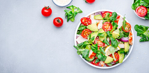Chicken salad with red tomato,  avocado, cucumber, red onion, lamb lettuce and sesame seeds on gray table background, top view banner