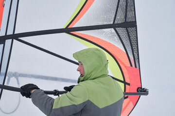 A middle-aged man, a snowsurfer, is making final preparations for riding a sailboard. Preparing for snowsurfing on a cloudy winter day.