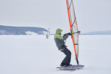 A middle-aged man, a snowsurfer, rides a sailboard on a snow-covered frozen lake. Snowsurfing on a cloudy winter day.