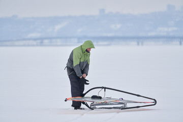 A middle-aged man, a snowsurfer, is making final preparations for riding a sailboard. Preparing for snowsurfing on a cloudy winter day.