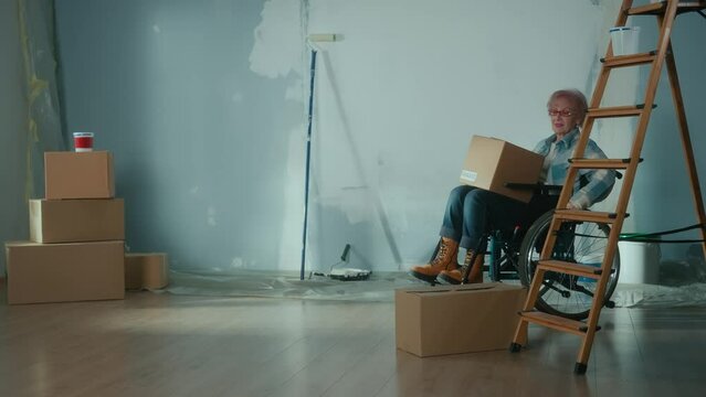 An elderly disabled woman moves in a wheelchair with a cardboard box. The granny puts the box looks around and plans repairs. Room with window, stepladder, wallpaper rolls, roller and paint.