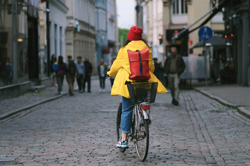 A young woman in a yellow windbreaker and a red winter hat rides a bicycle on a paved pedestrian path