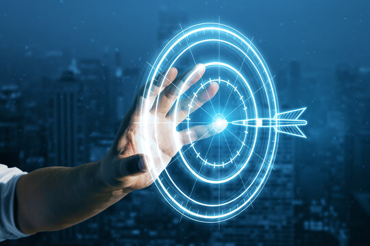 Target concept with a glowing hologram of bullseye and arrow hitting the center. Goals achievement, success. Close up of businessman hand using hud screen on blurry night city background.