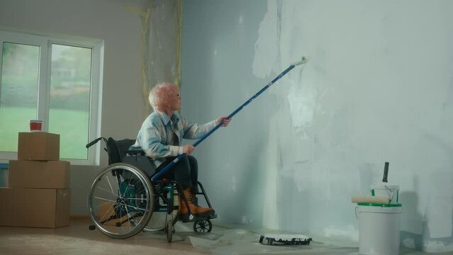 An elderly woman in a wheelchair paints a wall with white paint using a long roller. A disabled woman in the age makes repairs in the room. The concept of repair in the apartment.