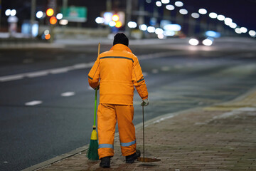Utility worker with broomstick and scoop for garbage work at night. Worker sweep city street with...