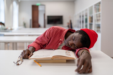 Exhausted African guy student fall asleep while studying in library, lying on desk with textbooks...