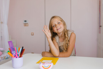 Caucasian girl is sitting at a desk with a notebook and stationery on it, raising her hand up at school